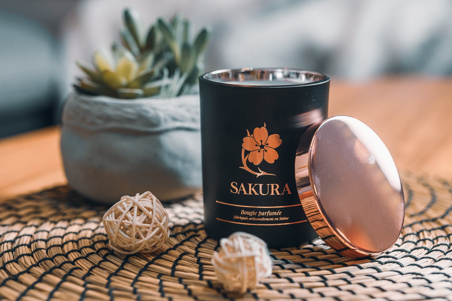 Bougie parfumée artisanale suisse Sakura en pot noir avec couvercle rosé, posée sur un set en rotin avec des décorations naturelles et une plante en arrière-plan – fabrication suisse.