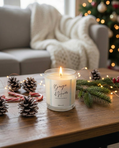 Bougie d’Hiver artisanale Sakura en cire de soja, allumée sur une table en bois dans une ambiance de Noël avec pommes de pin, branche de sapin et guirlandes lumineuses – fabrication suisse.