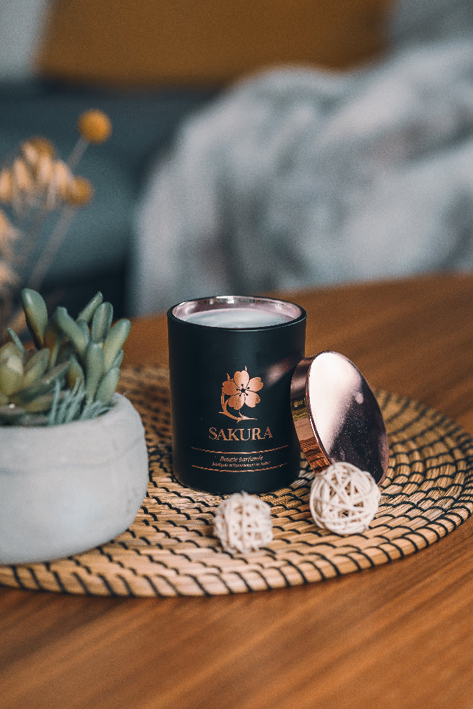 Bougie parfumée artisanale suisse Sakura en pot noir avec couvercle rosé, posée sur un set en rotin dans un intérieur cosy avec plante décorative – fabrication artisanale en Suisse.
