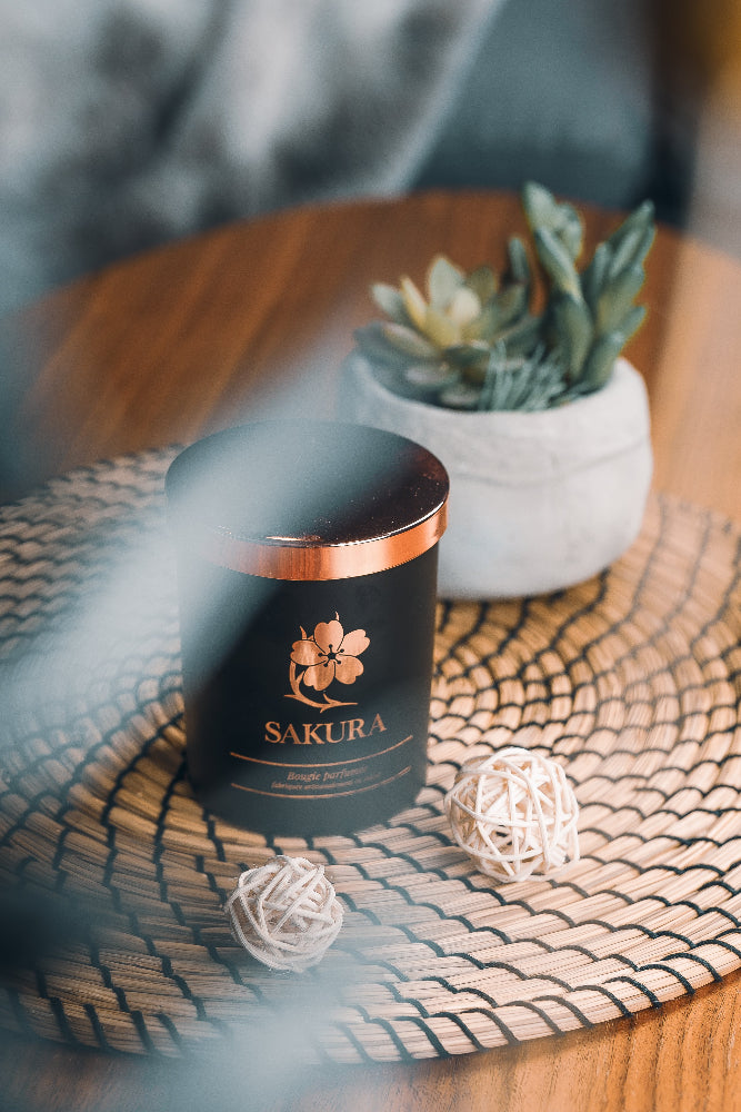 Bougie parfumée artisanale suisse Sakura en pot noir avec couvercle rosé, posée sur un set en rotin dans un intérieur cosy avec plante décorative – fabrication artisanale en Suisse.