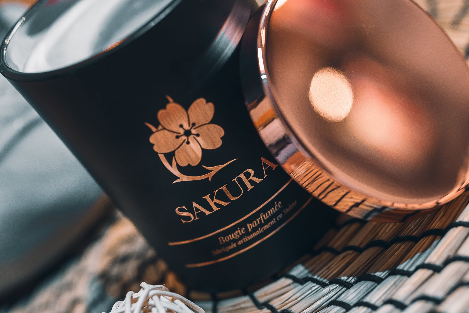 Bougie parfumée artisanale suisse Sakura en pot noir avec couvercle rosé, posée sur un set en rotin dans un intérieur cosy – fabrication artisanale en Suisse.