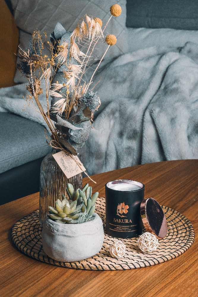 Bougie parfumée artisanale suisse Sakura en pot noir avec couvercle rosé, posée sur un set en rotin dans un intérieur cosy avec plante décorative – fabrication artisanale en Suisse.